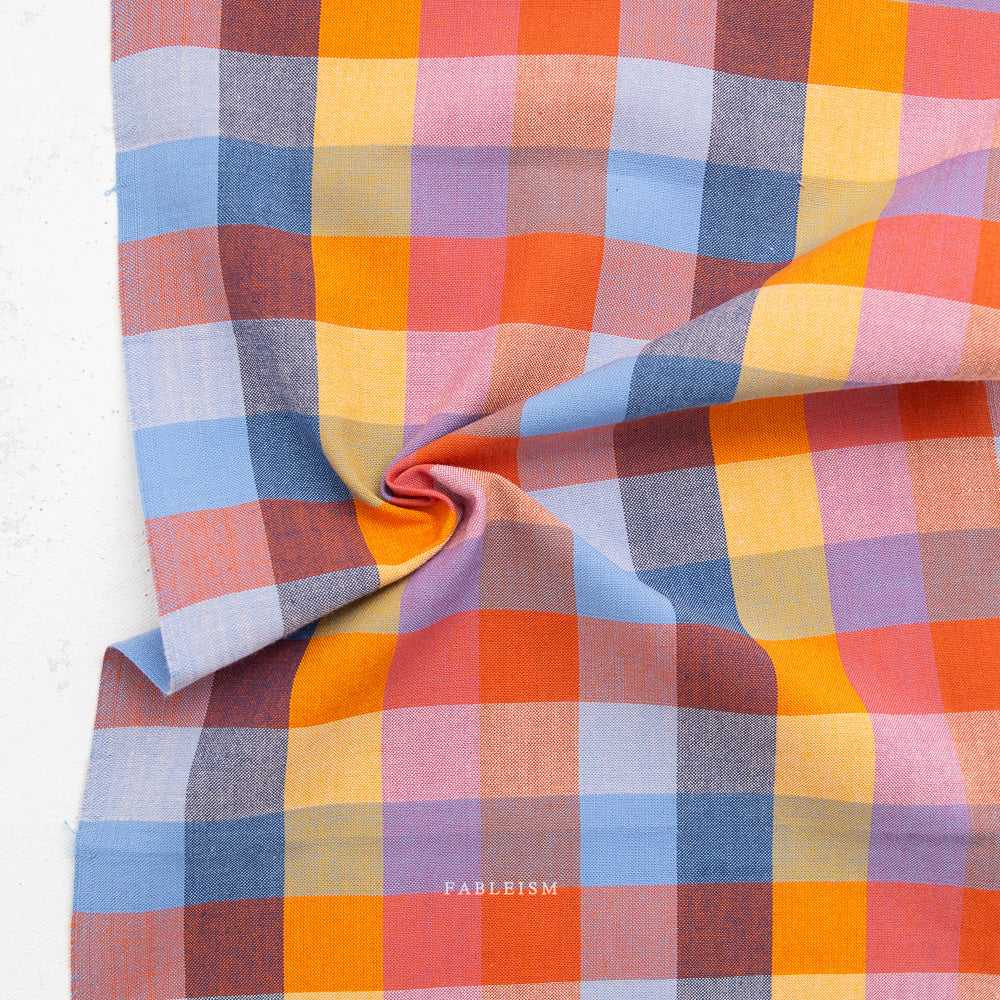 A close-up of a plaid fabric with a multicolor pattern of orange, pink, blue, and purple, draped to show its texture and pattern repeat. The brand name 'FABLEISM' is printed on the fabric.