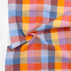 A close-up of a plaid fabric with a multicolor pattern of orange, pink, blue, and purple, draped to show its texture and pattern repeat. The brand name 'FABLEISM' is printed on the fabric.