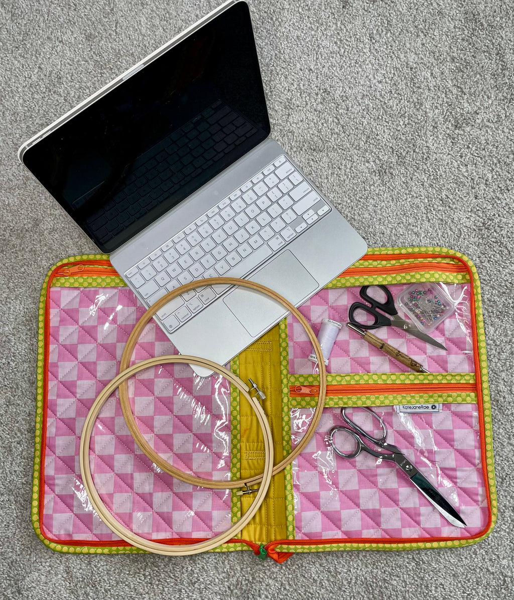 Laptop on a pink and yellow checkered mat with embroidery hoops and scissors.