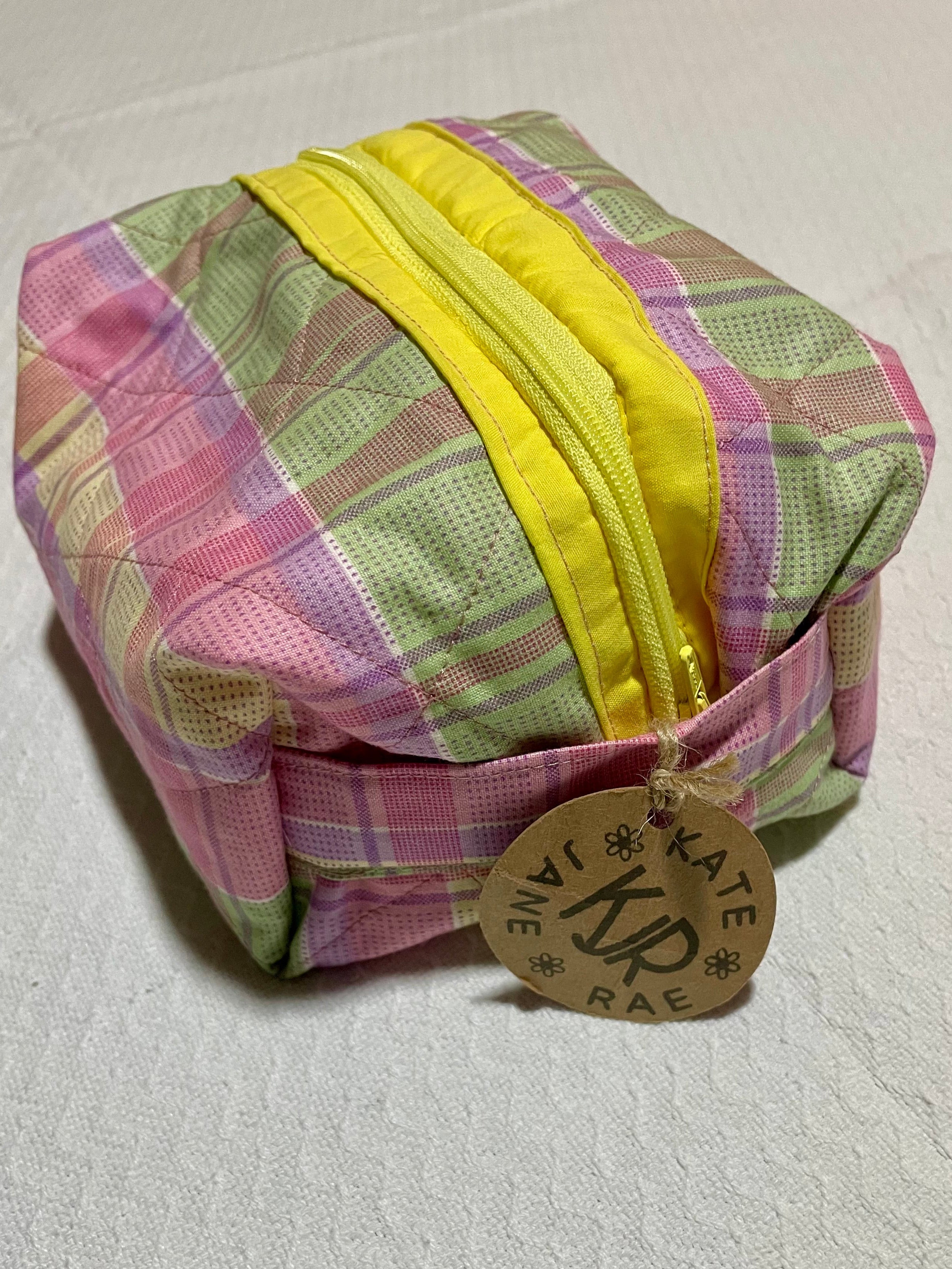 Colorful checkered fabric pouch with a yellow zipper on a textured white surface, featuring a brand tag.