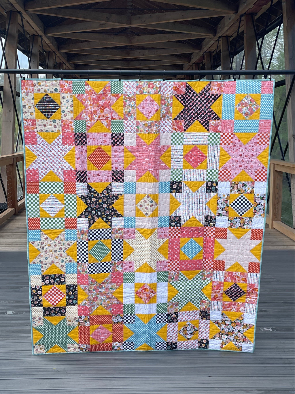 Colorful patchwork quilt displayed indoors with wooden beams and glass floor in the background