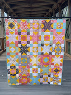 Colorful patchwork quilt displayed indoors with wooden beams and glass floor in the background