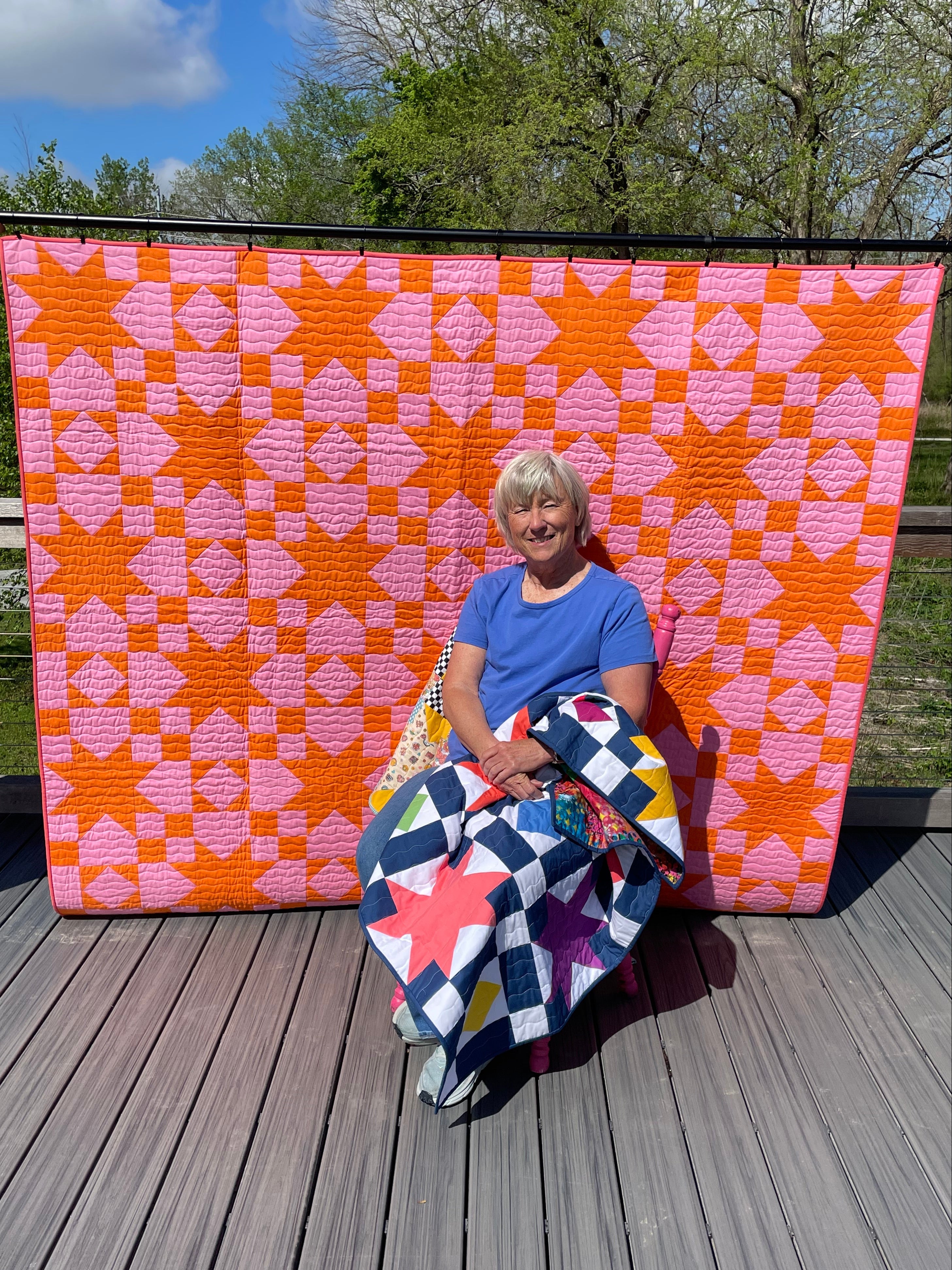 Person holding a colorful quilt on a wooden deck with trees in the background