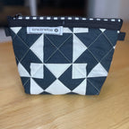 A small zippered pouch with a black and white geometric pattern, resting on a light wooden table. The pouch features a white brand label that reads 'Kate Jane Rose' with a small flower logo.