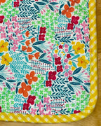 A square fabric zippered pouch or case with a colorful floral and geometric print, featuring a yellow gingham border, laid flat on a wooden surface.