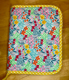 A square fabric-covered item, likely a notebook cover, pouch, or small case, featuring a colorful floral and geometric patchwork print with a yellow gingham border, placed on a wooden surface.