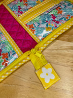 A close-up of a colorful zippered pouch with floral and quilted fabric panels, yellow trim, and a yellow flower pull tab, placed on a wooden surface.