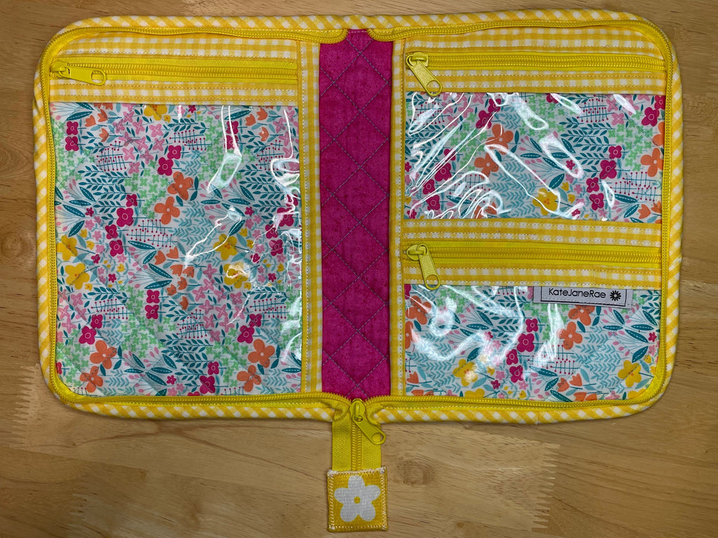 A quilted travel organizer pouch laid flat on a light wood floor. The pouch features a bright yellow and white gingham border, a central magenta quilted band, and three floral patterned pockets with clear plastic zippered inserts.