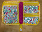 A quilted travel organizer pouch laid flat on a light wood floor. The pouch features a bright yellow and white gingham border, a central magenta quilted band, and three floral patterned pockets with clear plastic zippered inserts.