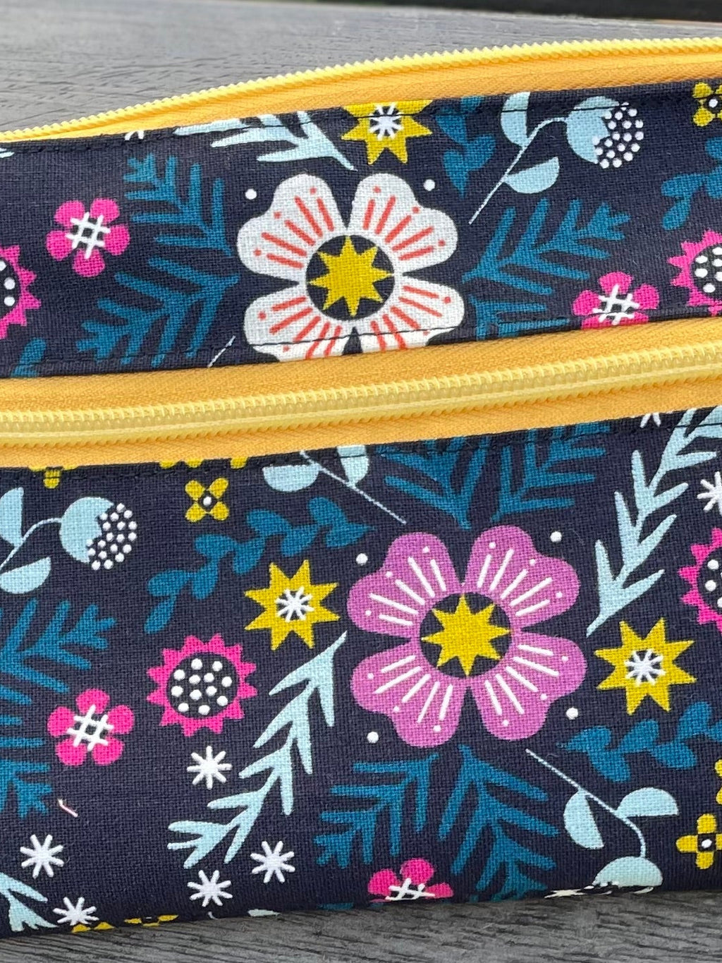 Floral-patterned pouch with a yellow zipper on a wooden surface with grass in the background