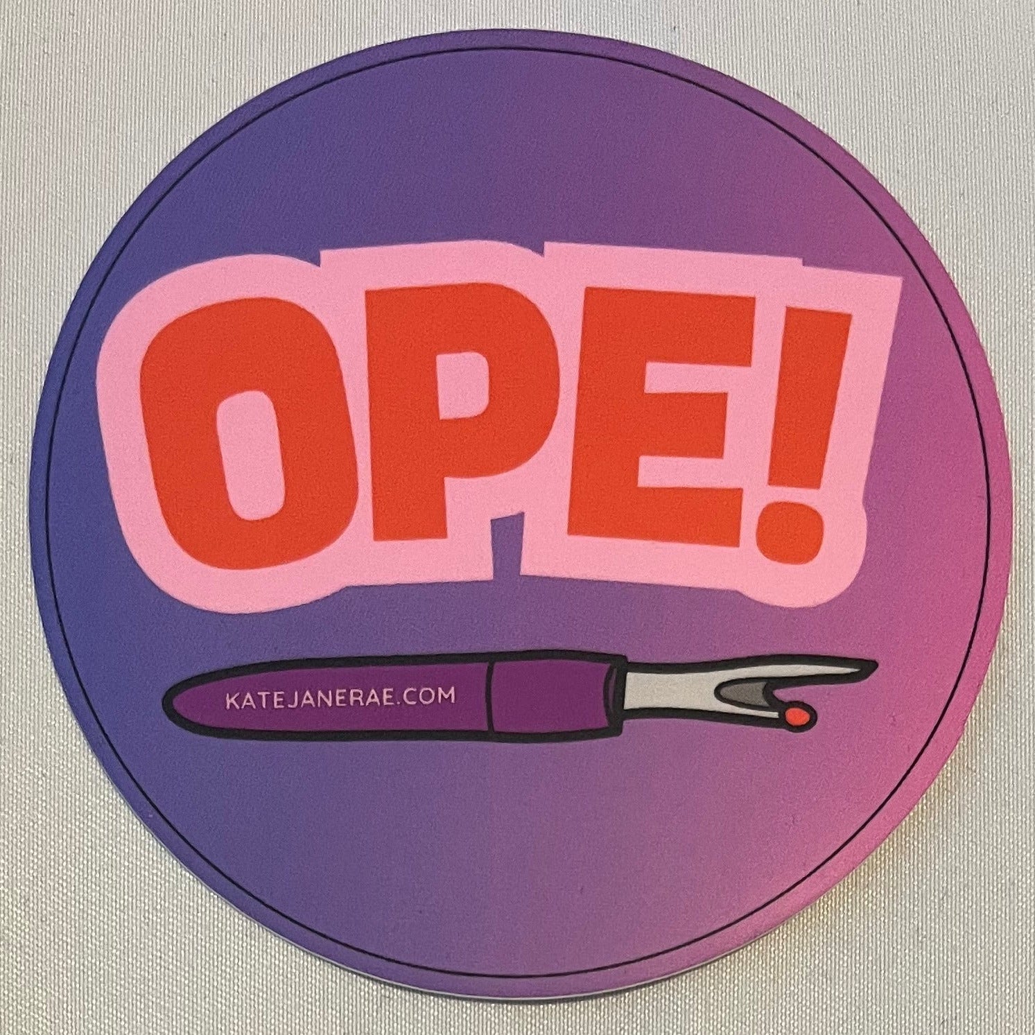 Circular sticker with 'OPE!' text and a pair of scissors on a beige background