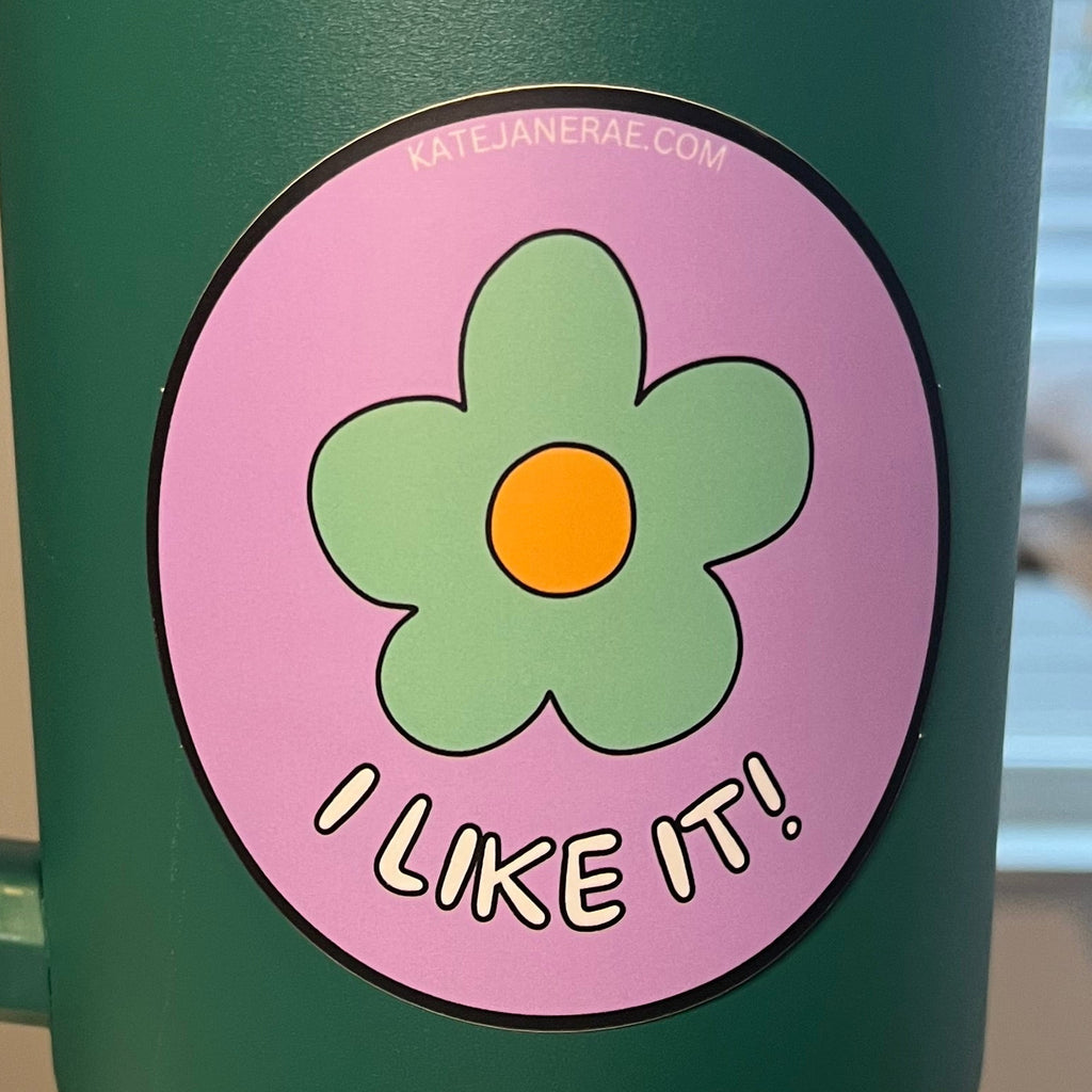 Green handle with a pink flower-shaped sticker labeled 'I Like It' on a blurred background.