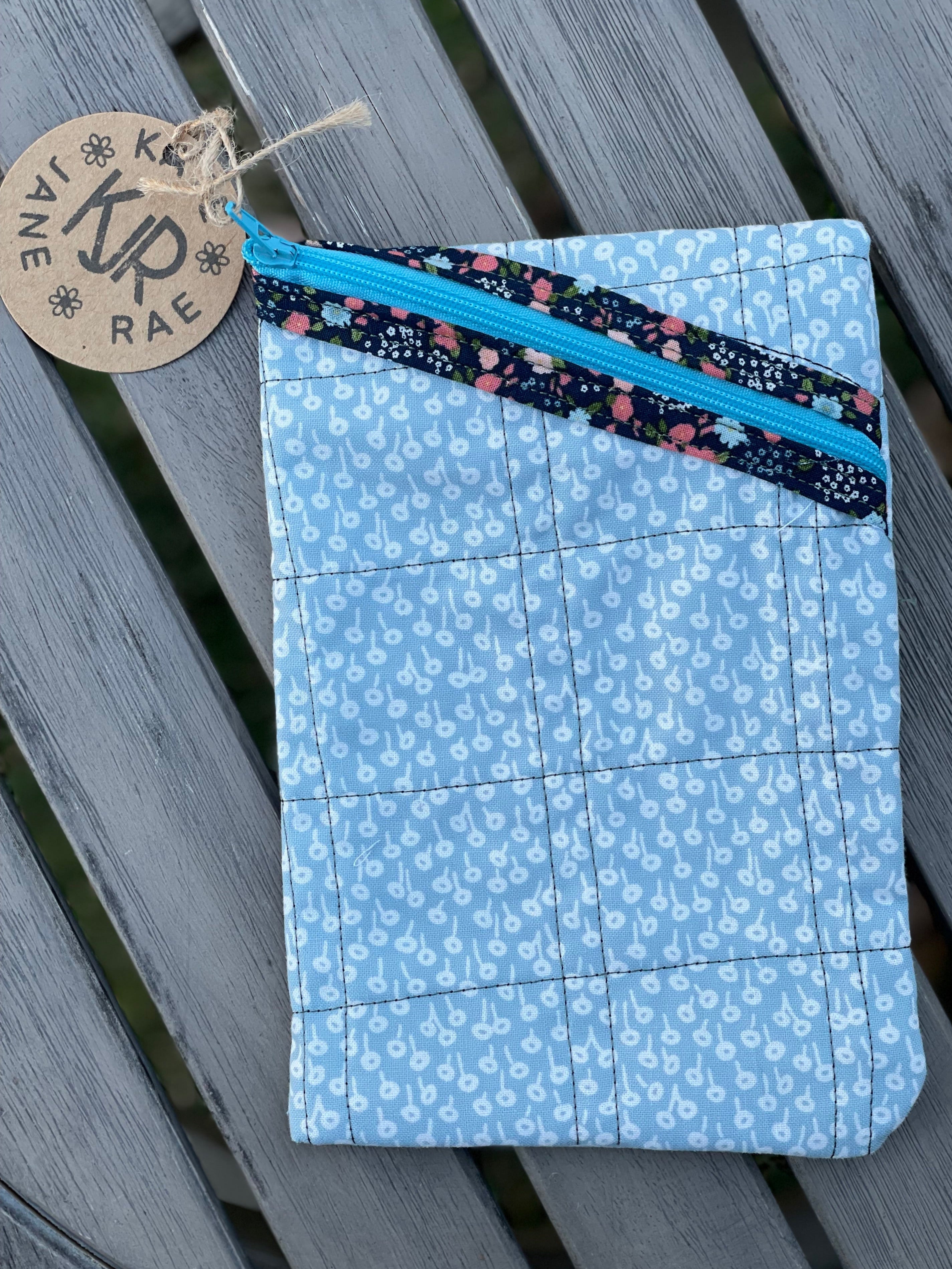 Blue fabric pouch with floral zipper tab on a wooden surface, featuring a brand tag.
