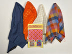Five folded fabric pieces in different patterns and colors are arranged on a white sheet, with a small booklet titled 'The Sampler quilt' placed on top. The fabrics include blue with white dots, orange with yellow motifs, cream and white gingham, and a multicolor plaid in blue, orange, yellow, and red.