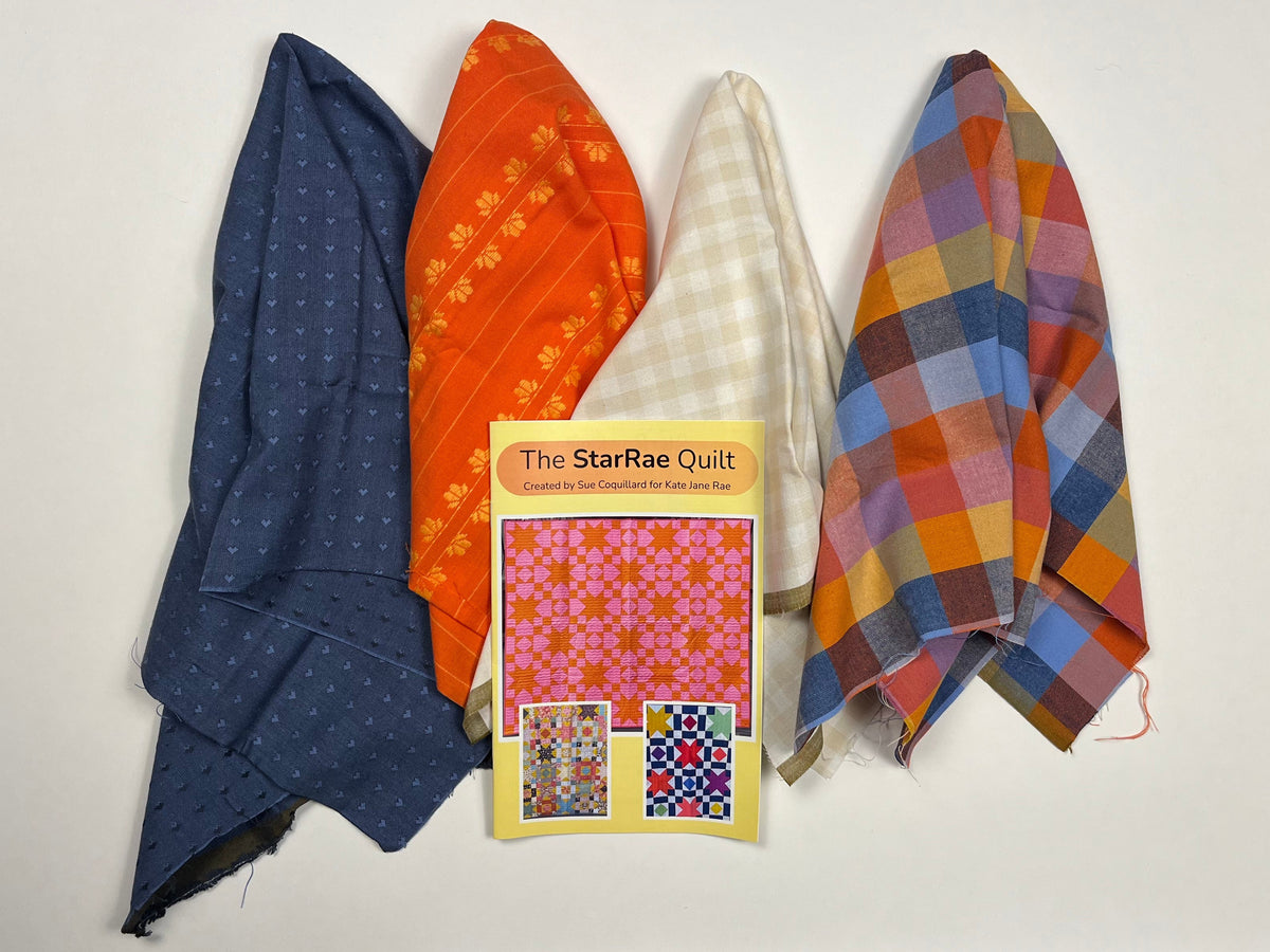 Five folded fabric pieces in different patterns and colors are arranged on a white sheet, with a small booklet titled 'The Sampler quilt' placed on top. The fabrics include blue with white dots, orange with yellow motifs, cream and white gingham, and a multicolor plaid in blue, orange, yellow, and red.