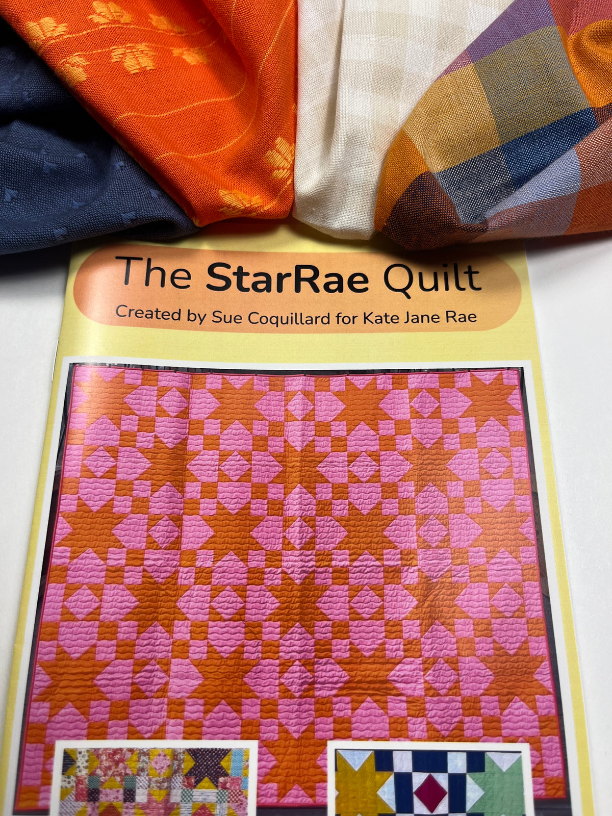 A quilt pattern booklet titled 'The StarRae Quilt' placed on top of several folded fabrics in orange, navy, cream, and plaid. The cover shows a pink and orange star quilt design.