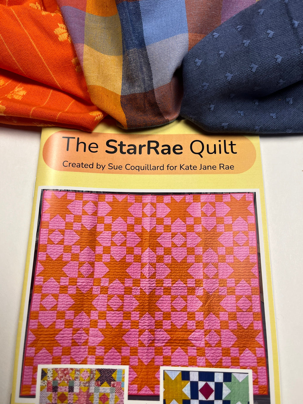 A quilt pattern booklet titled 'The StarRae Quilt' by Sue Coquillard for Kate Jane Rae, featuring a pink and orange star pattern cover, with folded colorful fabrics arranged above it on a white surface.