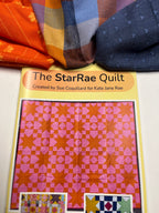 A quilt pattern booklet titled 'The StarRae Quilt' by Sue Coquillard for Kate Jane Rae, featuring a pink and orange star pattern cover, with folded colorful fabrics arranged above it on a white surface.