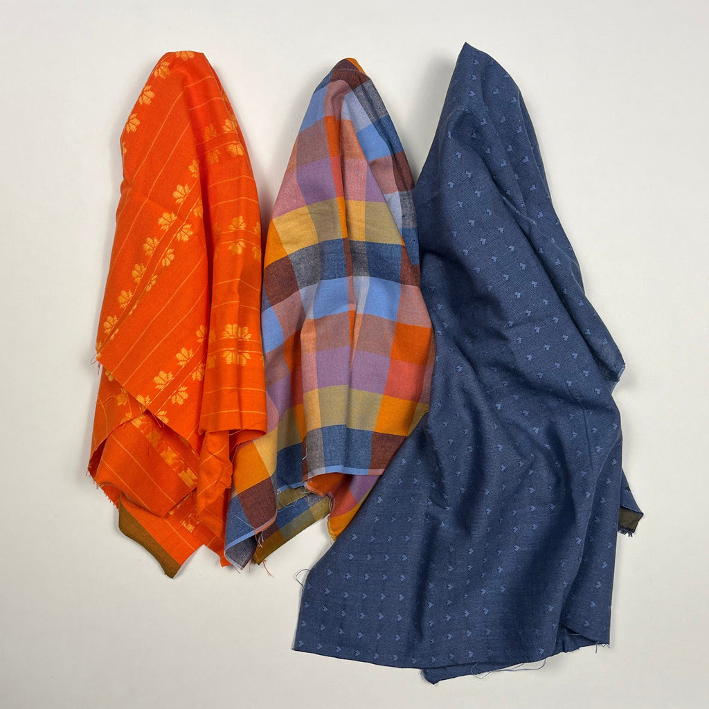 Three folded fabric items stacked on a white sheet: an orange patterned fabric at the top, a multicolor plaid fabric in the middle, and a navy blue polka-dot fabric at the bottom, photographed on a wooden surface.