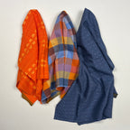 Three folded fabric items stacked on a white sheet: an orange patterned fabric at the top, a multicolor plaid fabric in the middle, and a navy blue polka-dot fabric at the bottom, photographed on a wooden surface.
