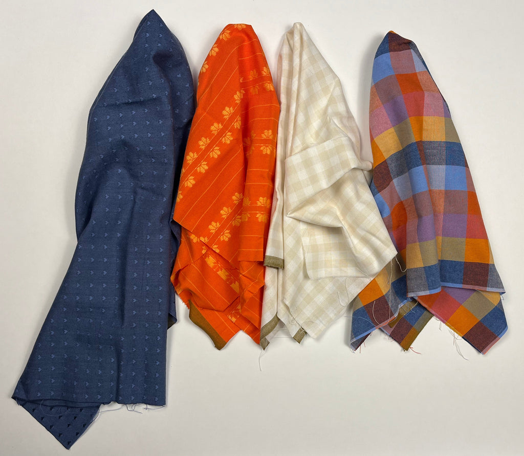 Five folded fabric pieces in different patterns and colors are stacked vertically on a white sheet. The fabrics include navy blue, orange, cream, and plaid patterns in blue, orange, red, and yellow.