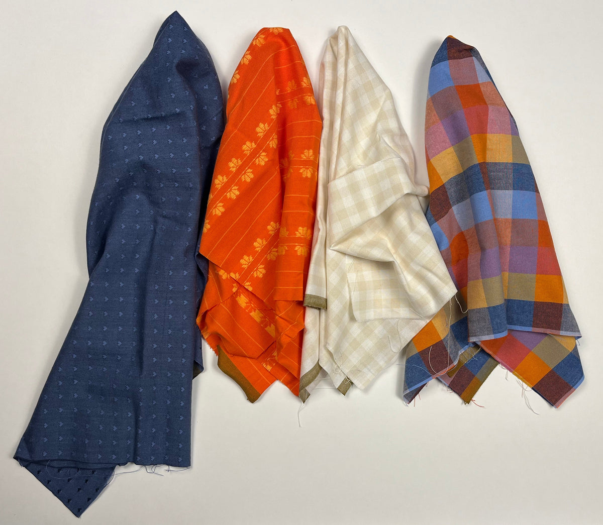 Five folded fabric pieces in different patterns and colors are stacked vertically on a white sheet. The fabrics include navy blue, orange, cream, and plaid patterns in blue, orange, red, and yellow.