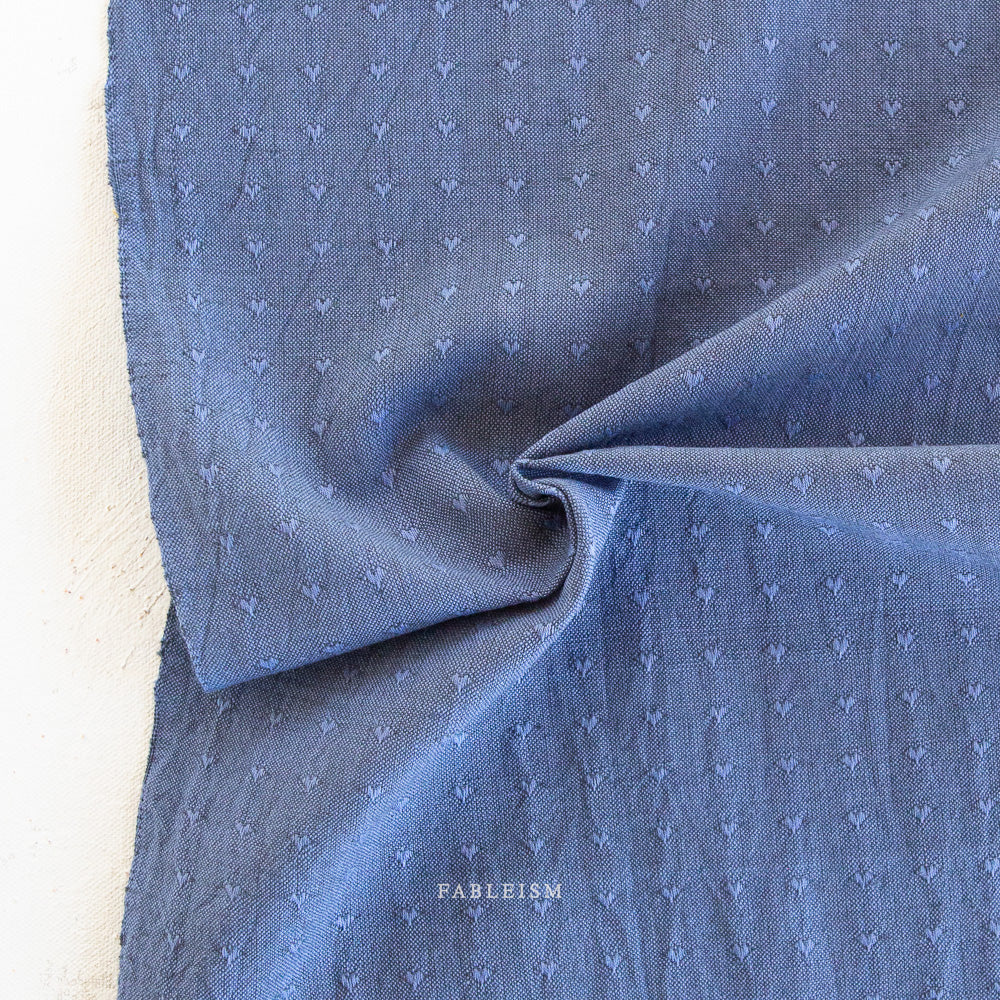 A close-up of blue fabric with a small repeating white embroidered or printed motif, draped to show texture and weave. A small visible brand mark reads 'FABLEISM' near the bottom center.
