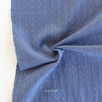 A close-up of blue fabric with a small repeating white embroidered or printed motif, draped to show texture and weave. A small visible brand mark reads 'FABLEISM' near the bottom center.