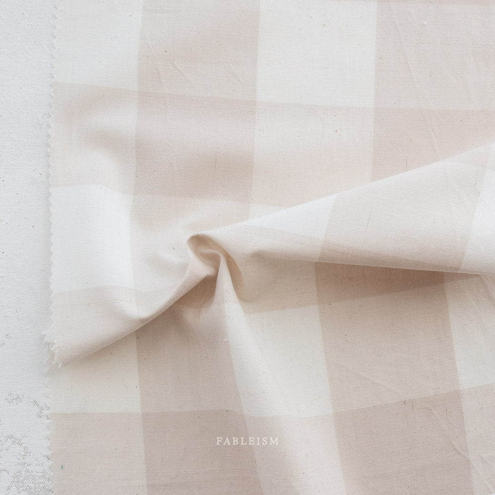 A close-up of a soft fabric with a pale blush pink and off-white gingham check pattern, draped to show its texture and weave.